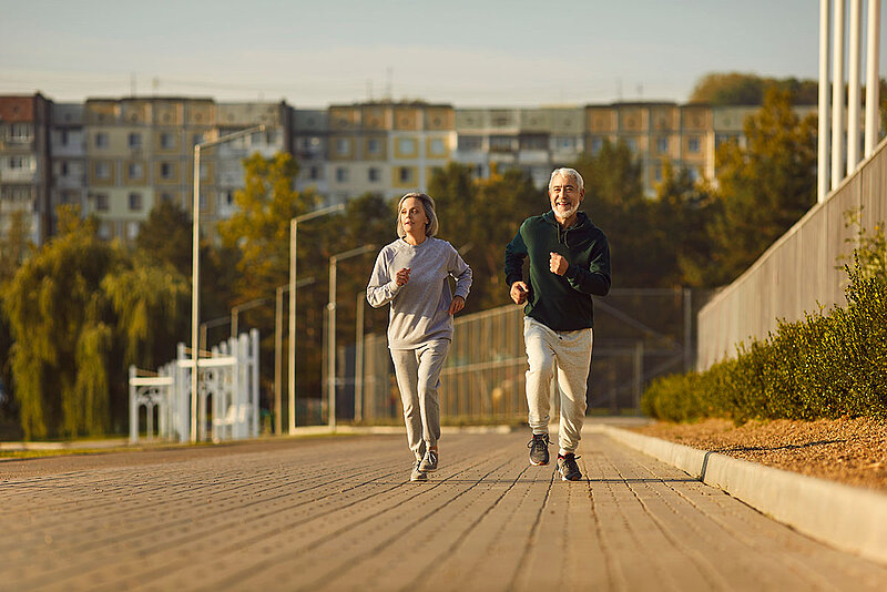 Bestager Paar joggt auf die Kamera zu, im Hintergrund eine Stadt