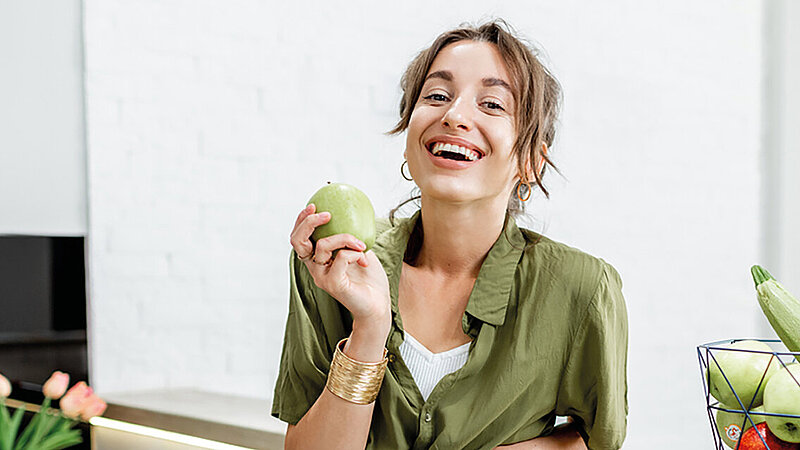In grün gekleidete Frau mit Mango in der Hand lacht fröhlich in die Kamera