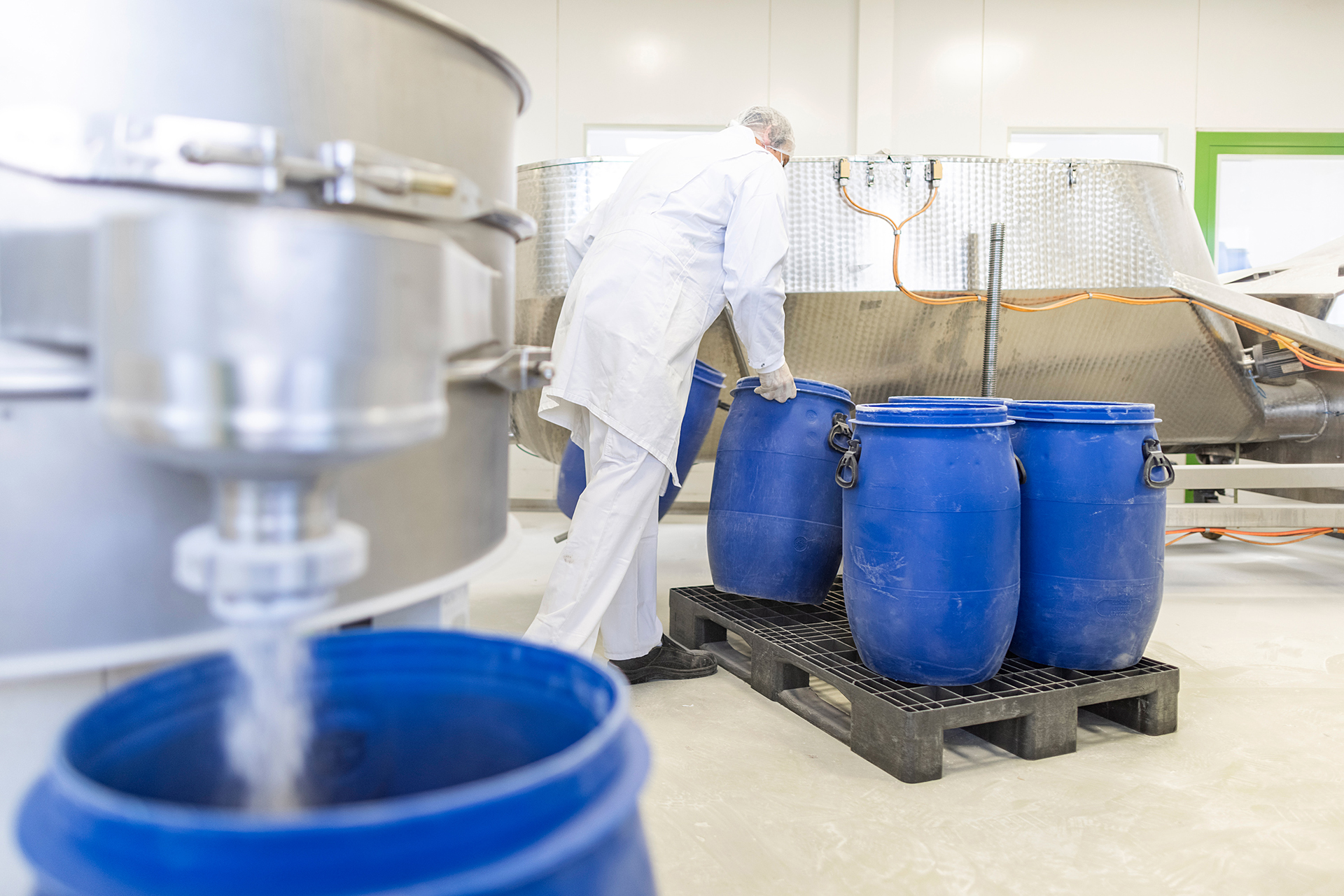 Im Vordergrund füllt eine Maschine Pulver in ein blaues Fass, im Hintergrung sammelt ein Mitarbeiter die bereits befüllten Fässer