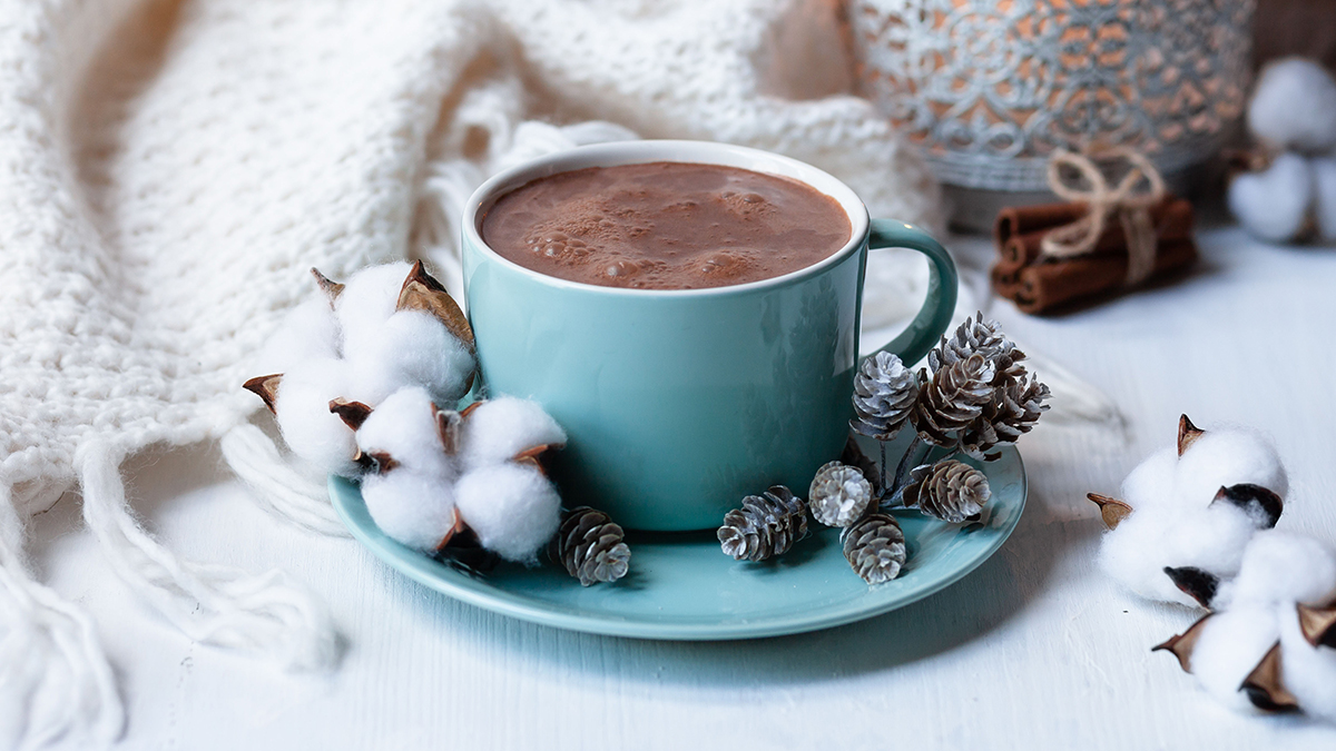 Blaue Tasse mit Kakao gefüllt, dazu winterliche Dekoration wie Schnee und Tannenzapfen