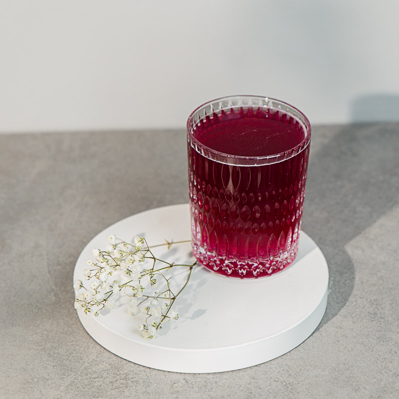 Roter Beauty Eistee in Kristallglas auf einem weißen Tablett, daneben Schleierkraut