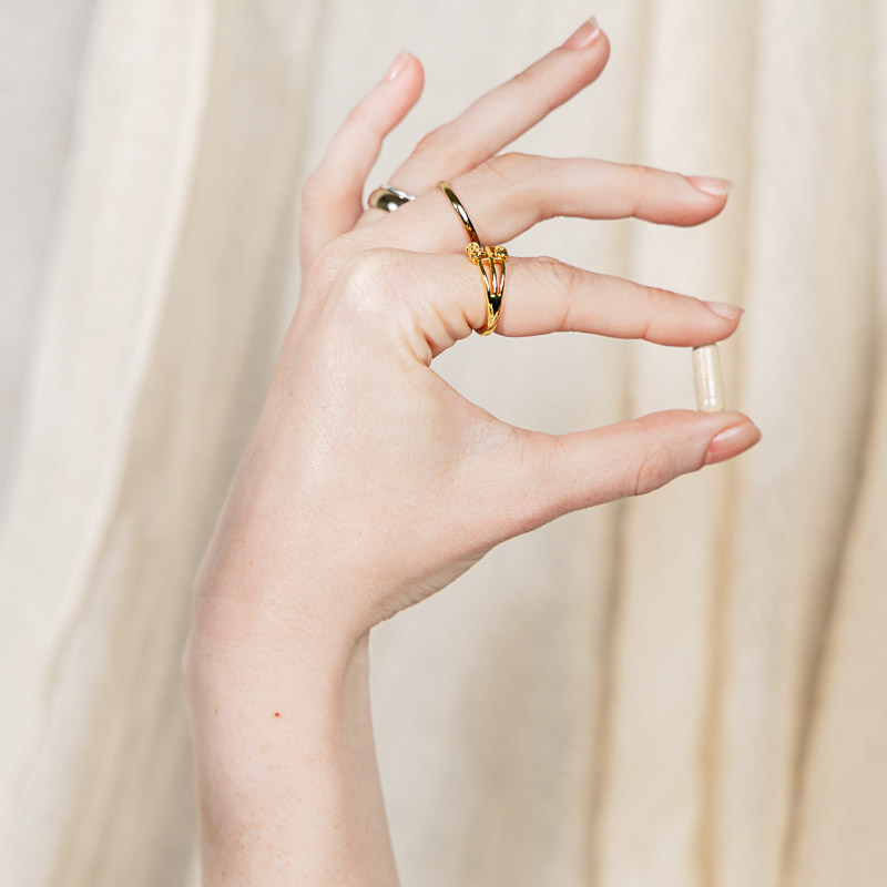 Eine Hand mit goldenen Ringen hält eine Kapsel zwischen zwei Fingern. Im Hintergrund beiger Stoff