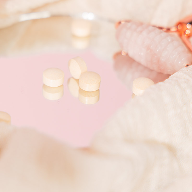 Auf einer spiegelnden Oberfläche liegen kleine runde Tabletten. Daneben ein Rosenquarzroller umgeben von weichem Stoff
