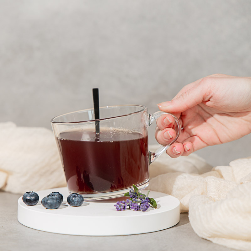 Frauengesundheit-Tee in einer Glastasse, die von einer Hand leicht angehoben wird,dazu Heidelbeeren und Lavendel