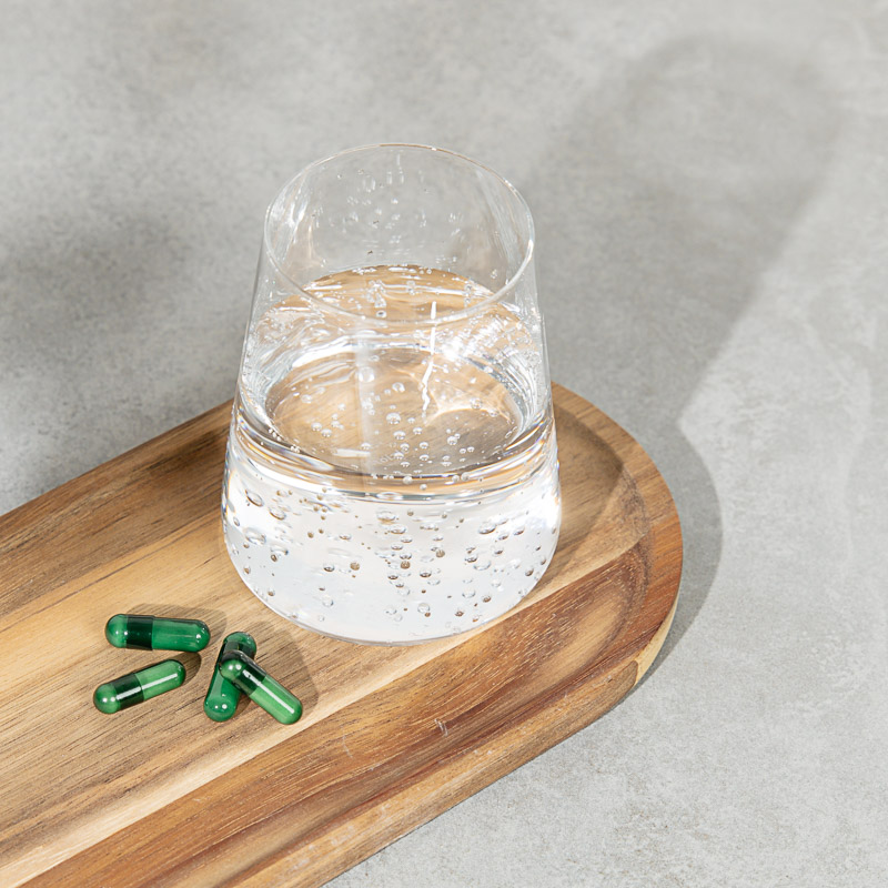 Kapseln für das Herz Kreislauf System liegen auf einem Holzbrett, dazu ein glas Mineralwasser