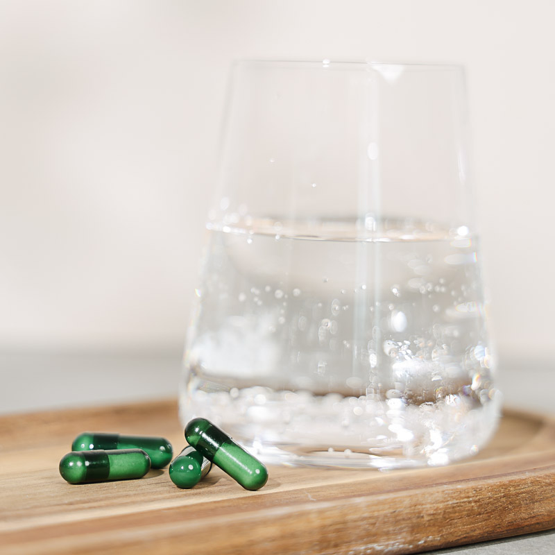 Kapseln für das Herz Kreislauf System liegen auf einem Holzbrett, dazu ein glas Mineralwasser