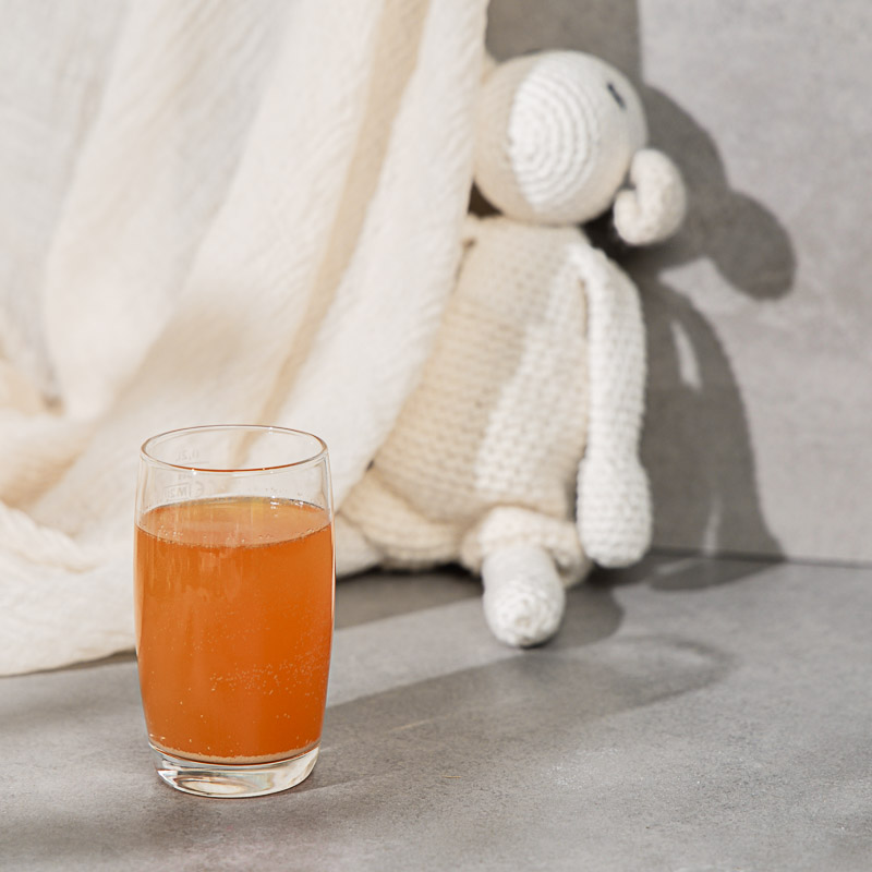 Orangener Nahrungsergänzungsmittel Drink für Kinder in einem Glas. Im Hintergrund sitzt ein Kuscheltier
