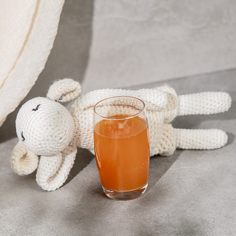 Orangener Nahrungsergänzungsmittel Drink für Kinder in einem Glas. Im Hintergrund liegt ein Kuscheltier