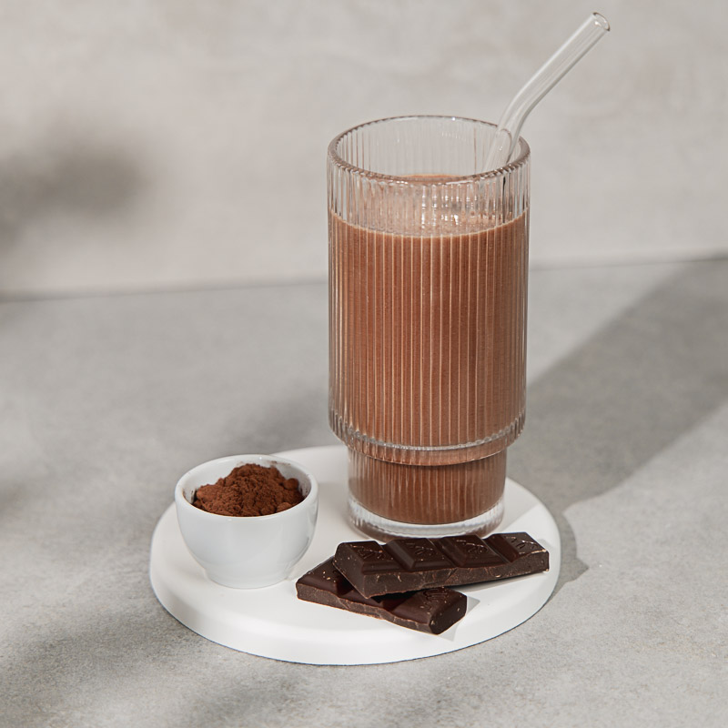 Mahlzeitenersatz Shake Schoko in einem Hohen Glas mit Strohhalm, dazu Schokolade und Pulver zum anmischen des Shakes daneben