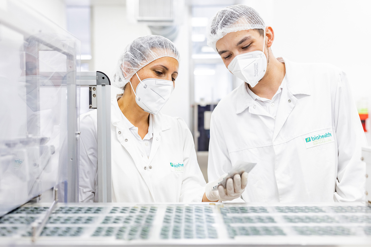 Mitarbeiterin aus der Produktion erklärt einem Quereinsteiger seinen Arbeitsplatz. Beide tragen weiße Produktionskleidung mit Masken und Haarnetzen
